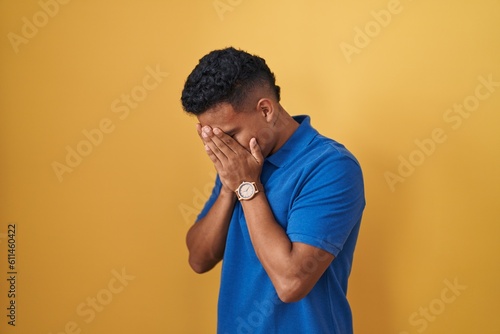 Young hispanic man standing over yellow background with sad expression covering face with hands while crying. depression concept.