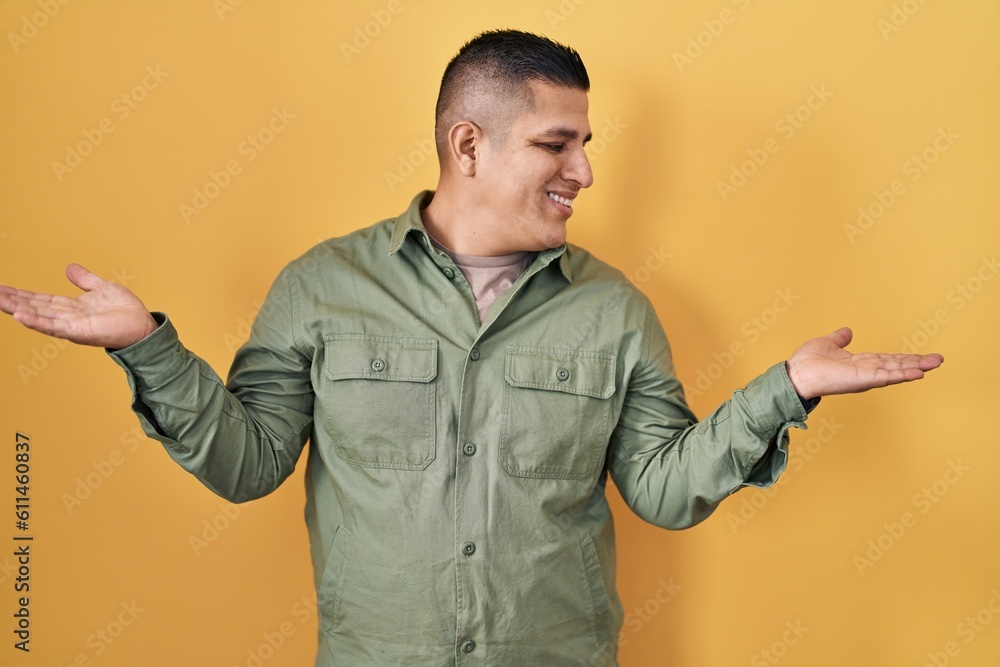 Hispanic young man standing over yellow background smiling showing both hands open palms, presenting and advertising comparison and balance Hispanic young man standing over yellow background smiling showing both hands open palms, presenting and advertising comparison and balance