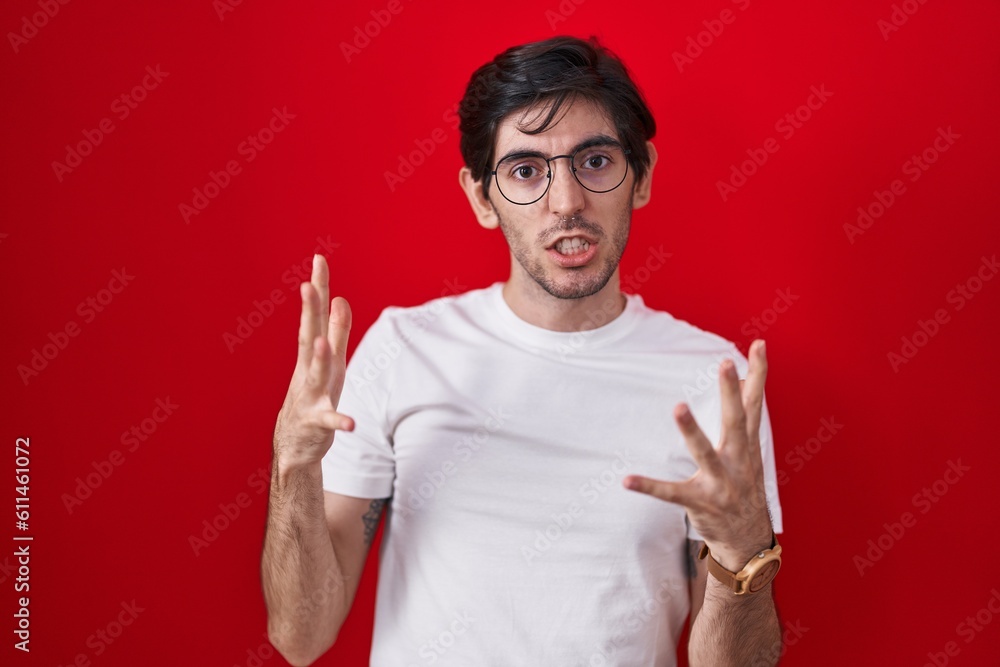 Naklejka premium Young hispanic man standing over red background crazy and mad shouting and yelling with aggressive expression and arms raised. frustration concept.