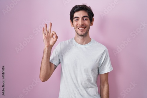 Young hispanic man standing over pink background smiling positive doing ok sign with hand and fingers. successful expression.