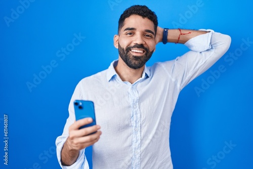 Hispanic man with beard using smartphone typing message smiling confident touching hair with hand up gesture, posing attractive and fashionable