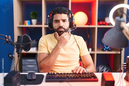 Hispanic man with beard playing video games with headphones looking confident at the camera with smile with crossed arms and hand raised on chin. thinking positive.