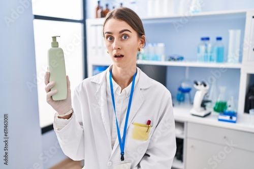 Young caucasian woman working at scientist laboratory holding body lotion scared and amazed with open mouth for surprise  disbelief face