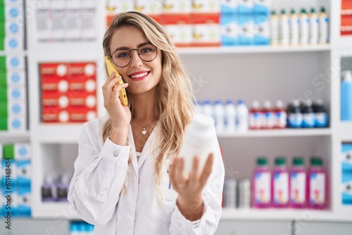 Young woman pharmacist talking on smartphone holding pills bottle at pharmacy