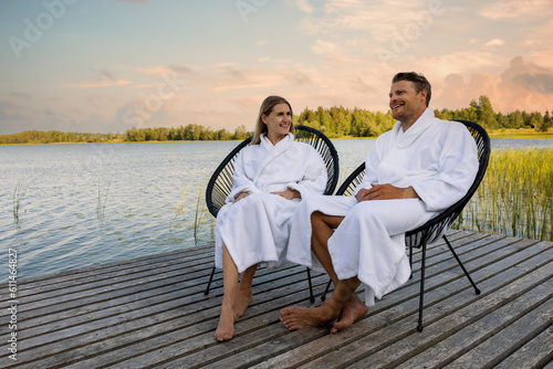 happy couple in white bathrobes relaxing on wooden lake footbridge after spa treatments at warm summer evening photo