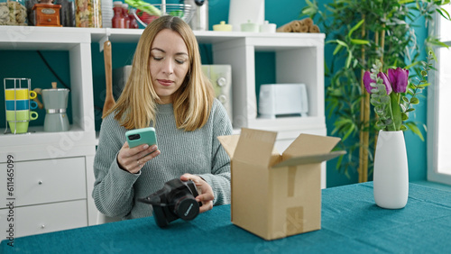 Young blonde woman unpacking professional camera of cardboard using smartphone at dinning room © Krakenimages.com