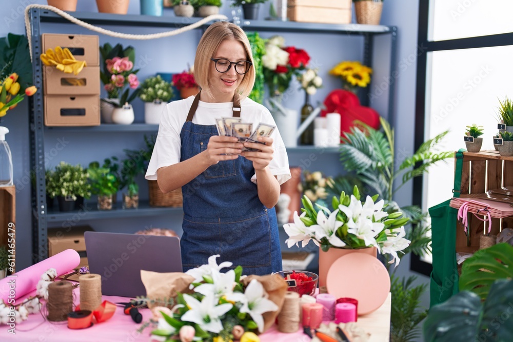 Young blonde woman florist using laptop counting dollars at florist shop