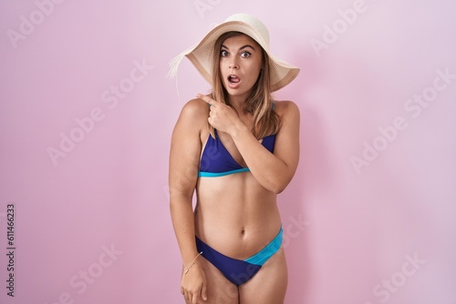 Young hispanic woman wearing bikini over pink background surprised pointing with finger to the side, open mouth amazed expression.