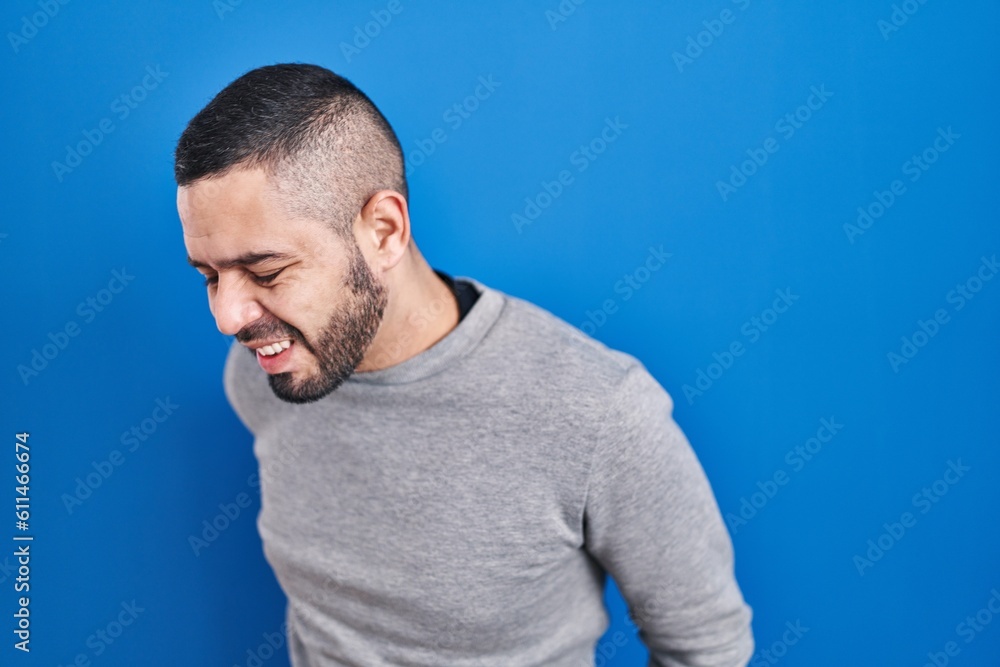 Hispanic man standing over blue background suffering of backache, touching back with hand, muscular pain Hispanic man standing over blue background suffering of backache, touching back with hand, muscular pain
