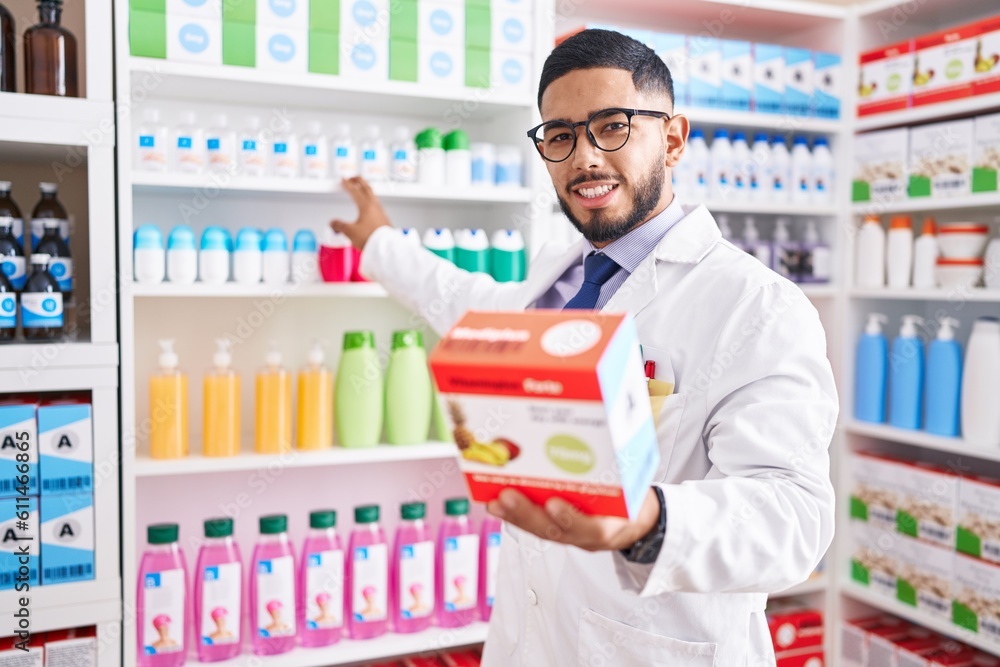 Young latin man pharmacist smiling confident holding vitamin package at pharmacy