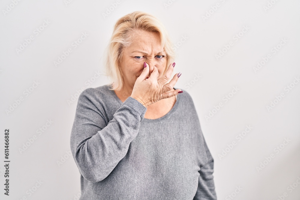 Middle age caucasian woman standing over white background smelling something stinky and disgusting, intolerable smell, holding breath with fingers on nose. bad smell Middle age caucasian woman standing over white background smelling something stinky and disgusting, intolerable smell, holding breath with fingers on nose. bad smell