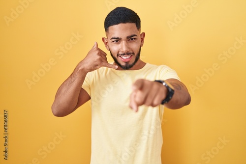 Young hispanic man standing over yellow background smiling doing talking on the telephone gesture and pointing to you. call me.