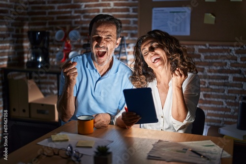 Middle age hispanic couple using touchpad sitting on the table at night very happy and excited doing winner gesture with arms raised, smiling and screaming for success. celebration concept.