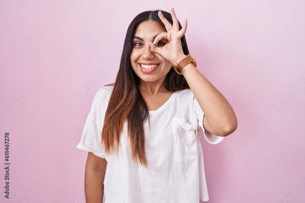 Young arab woman standing over pink background doing ok gesture with hand smiling, eye looking through fingers with happy face. Young arab woman standing over pink background doing ok gesture with hand smiling, eye looking through fingers with happy face.