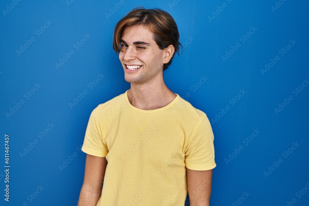 Young man standing over blue background winking looking at the camera with sexy expression, cheerful and happy face.