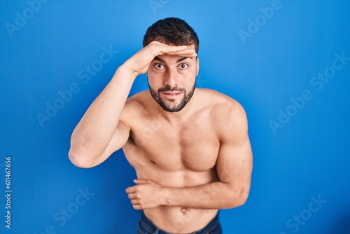 Handsome hispanic man standing shirtless very happy and smiling looking far away with hand over head. searching concept.