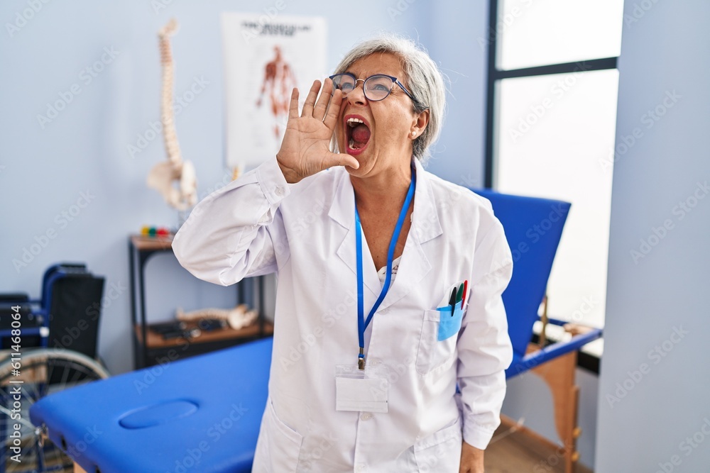 Middle age woman with grey hair working at pain recovery clinic shouting and screaming loud to side with hand on mouth. communication concept. Middle age woman with grey hair working at pain recovery clinic shouting and screaming loud to side with hand on mouth. communication concept.