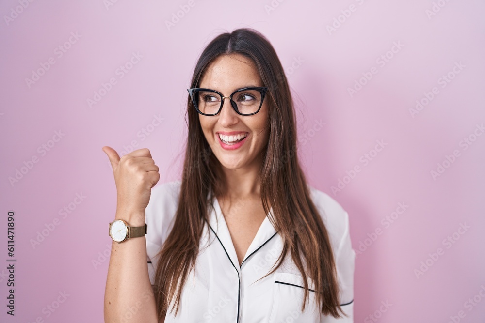 Young brunette woman wearing glasses standing over pink background smiling with happy face looking and pointing to the side with thumb up. Young brunette woman wearing glasses standing over pink background smiling with happy face looking and pointing to the side with thumb up.