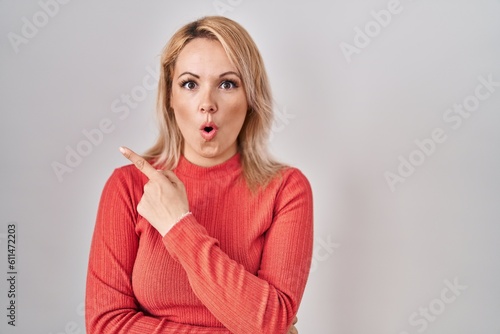 Blonde woman standing over isolated background surprised pointing with finger to the side, open mouth amazed expression. © Krakenimages.com