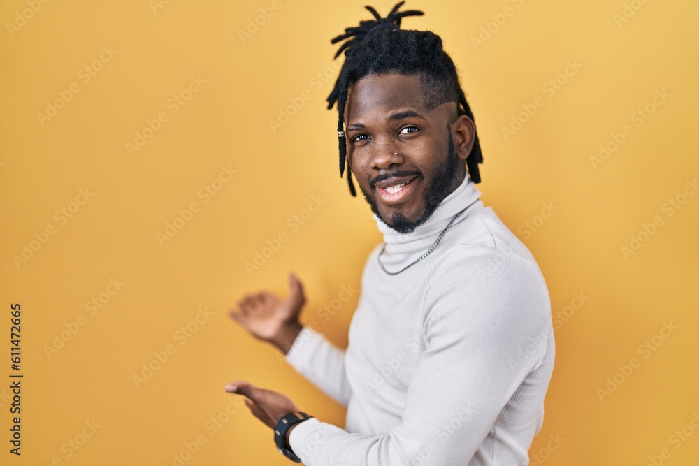 African man with dreadlocks wearing turtleneck sweater over yellow background inviting to enter smiling natural with open hand African man with dreadlocks wearing turtleneck sweater over yellow background inviting to enter smiling natural with open hand