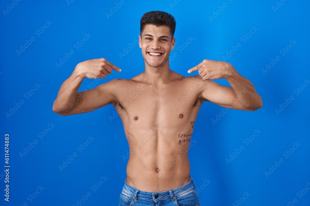 Young hispanic man standing shirtless over blue background looking confident with smile on face, pointing oneself with fingers proud and happy.