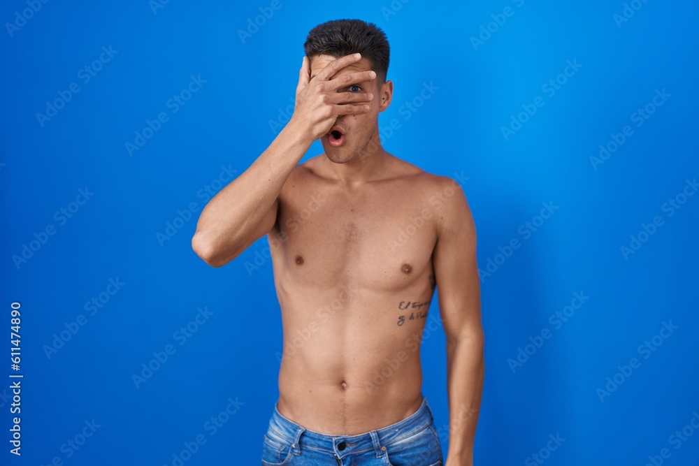 Fototapeta premium Young hispanic man standing shirtless over blue background peeking in shock covering face and eyes with hand, looking through fingers with embarrassed expression.