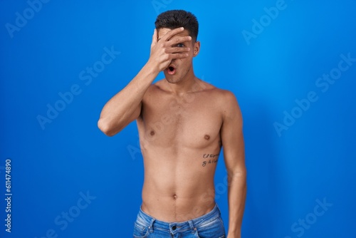 Young hispanic man standing shirtless over blue background peeking in shock covering face and eyes with hand, looking through fingers with embarrassed expression.