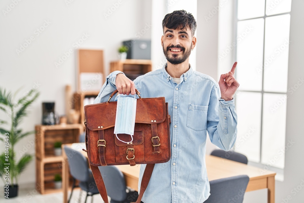 Young hispanic man with beard working at the office holding suitcase and safety mask smiling happy pointing with hand and finger to the side Young hispanic man with beard working at the office holding suitcase and safety mask smiling happy pointing with hand and finger to the side