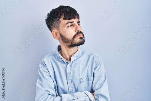 Young hispanic man with beard standing over blue background looking to the side with arms crossed convinced and confident