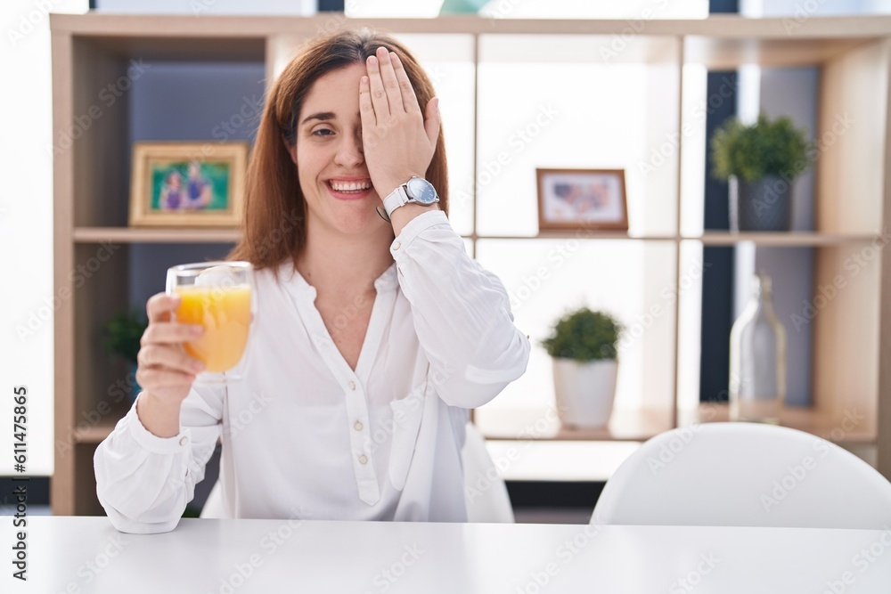 Brunette woman drinking glass of orange juice covering one eye with hand, confident smile on face and surprise emotion. Brunette woman drinking glass of orange juice covering one eye with hand, confident smile on face and surprise emotion.