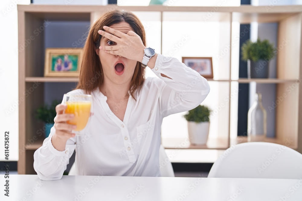 Brunette woman drinking glass of orange juice peeking in shock covering face and eyes with hand, looking through fingers with embarrassed expression. Brunette woman drinking glass of orange juice peeking in shock covering face and eyes with hand, looking through fingers with embarrassed expression.