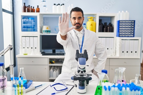 Young hispanic man with beard working at scientist laboratory doing stop sing with palm of the hand. warning expression with negative and serious gesture on the face.
