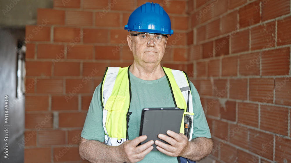 Middle age grey-haired man builder holding touchpad with relaxed expression at construction site Middle age grey-haired man builder holding touchpad with relaxed expression at construction site
