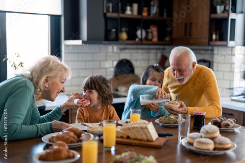 Grandchildren having breakfast with grandparents in home atmosphere. Cute small boy eating donut while grandmother is feeding him. Cheerful little girl preparing cereals with grandfather.