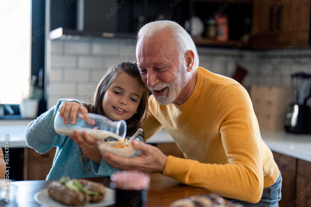 Smiling little girl with affectionate senior grandfather preparing breakfast in the kitchen at home, adding milk into bowl with cereals. Granddaughter and grandfather having cornflakes in the morning. Smiling little girl with affectionate senior grandfather preparing breakfast in the kitchen at home, adding milk into bowl with cereals. Granddaughter and grandfather having cornflakes in the morning.