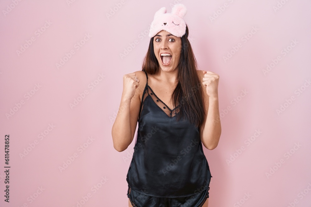 Young brunette woman wearing sleep mask and pyjama celebrating surprised and amazed for success with arms raised and open eyes. winner concept. Young brunette woman wearing sleep mask and pyjama celebrating surprised and amazed for success with arms raised and open eyes. winner concept.