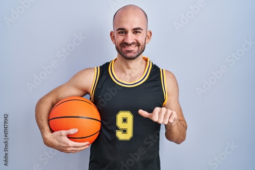 Young bald man with beard wearing basketball uniform holding ball pointing to the back behind with hand and thumbs up, smiling confident