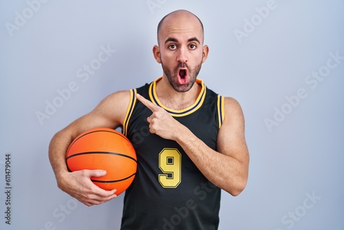 Young bald man with beard wearing basketball uniform holding ball surprised pointing with finger to the side, open mouth amazed expression.