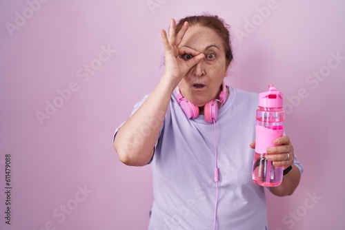 Senior woman wearing sportswear and headphones doing ok gesture shocked with surprised face, eye looking through fingers. unbelieving expression.
