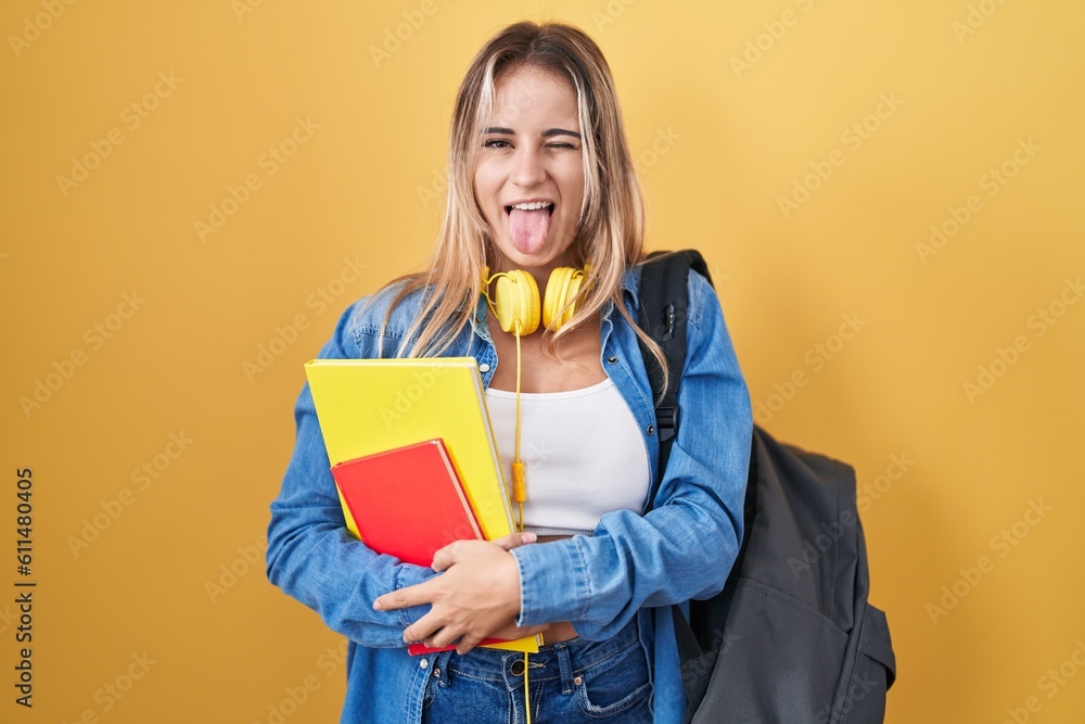 Young blonde woman wearing student backpack and holding books sticking tongue out happy with funny expression. emotion concept. Young blonde woman wearing student backpack and holding books sticking tongue out happy with funny expression. emotion concept.