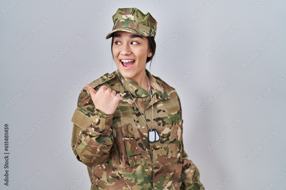 Young south asian woman wearing camouflage army uniform smiling with happy face looking and pointing to the side with thumb up.