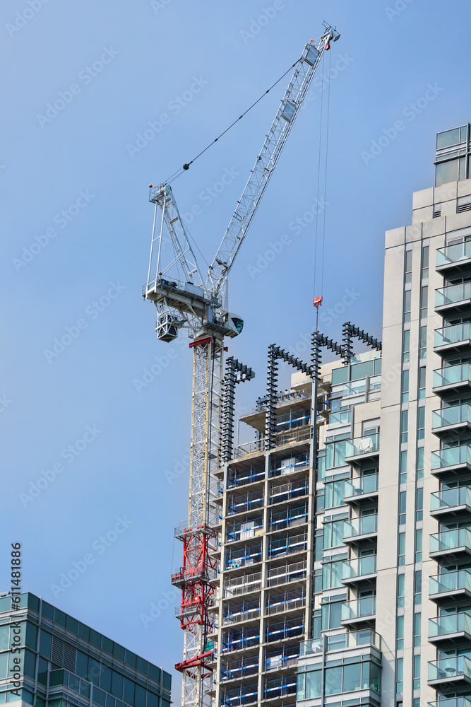 Fototapeta premium Large industrial crane machine working near tall skyscraper building, clear sky background. Modern construction industry concept