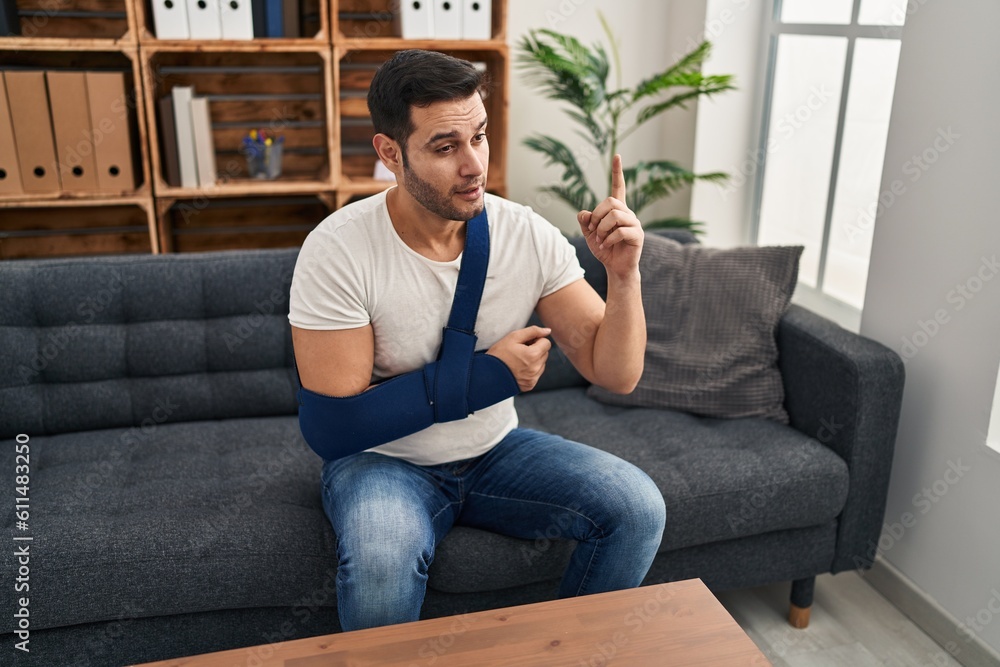 Young hispanic man with beard wearing arm on sling sitting at therapy consult surprised with an idea or question pointing finger with happy face, number one