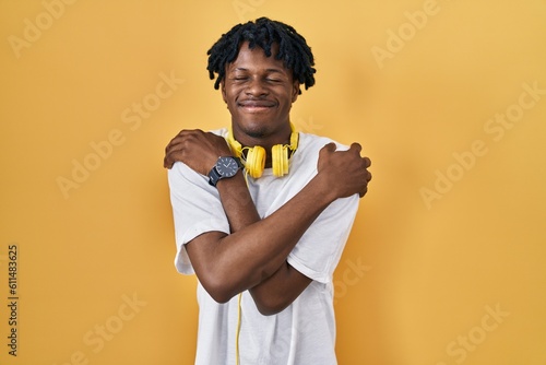 Canvas-taulu Young african man with dreadlocks standing over yellow background hugging oneself happy and positive, smiling confident