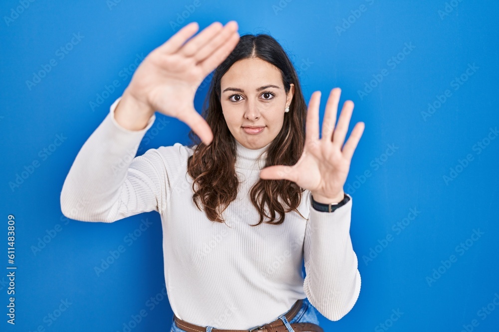 Young hispanic woman standing over blue background doing frame using hands palms and fingers, camera perspective Young hispanic woman standing over blue background doing frame using hands palms and fingers, camera perspective