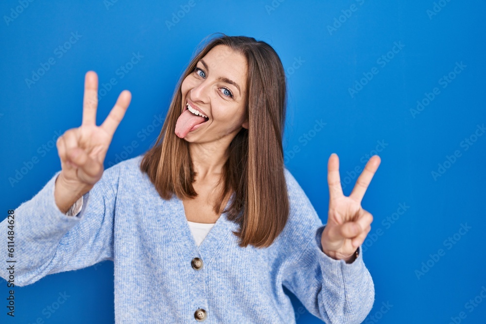 Young woman standing over blue background smiling with tongue out showing fingers of both hands doing victory sign. number two. Young woman standing over blue background smiling with tongue out showing fingers of both hands doing victory sign. number two.