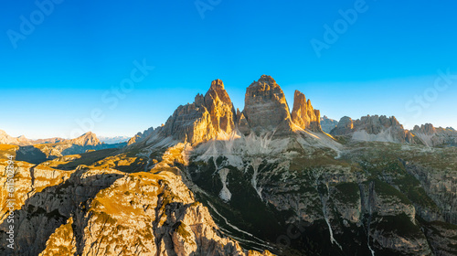 Sun rises over mountain range. High rocky peaks with sand-covered foothills of Three Peaks of Lavaredo under cloudless sky aerial view in back lit photo