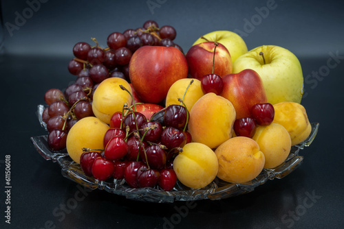 A plate of fruits consisting of grapes  peaches  apricots  cherries and apples on a black background