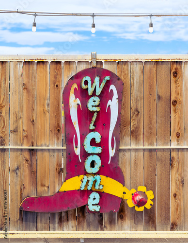 A red, yellow and turquoise Welcome Sign  in the shape of a cowboy boot attached to a high wooden slat fence.