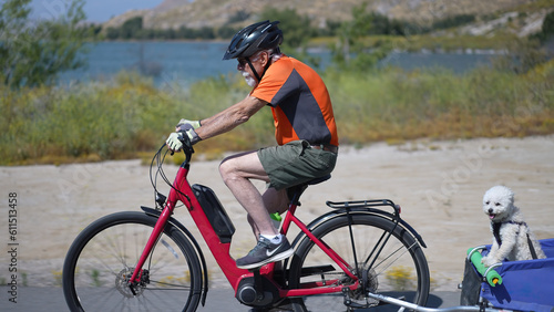 Elderly senior man biking on an e-bike on a trail pulling a trailer with a dog in it.